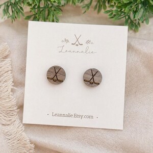 May include: Wooden stud earrings featuring a hockey stick design. The earrings are round with a dark wood grain finish. The earrings are displayed on a white card with the text "Leannalie.Etsy.com".