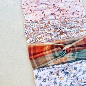 May include: Five different patterned headbands stacked on top of each other. The top headband is pink with white flowers and brown leaves. The second headband is pink with a brown and white donut, coffee cup, and pumpkin pattern. The third headband is a plaid pattern with red, green, and brown squares. The fourth headband is white with a light pink pumpkin, coffee cup, and smiley face pattern. The bottom headband is white with a pink pumpkin pattern.