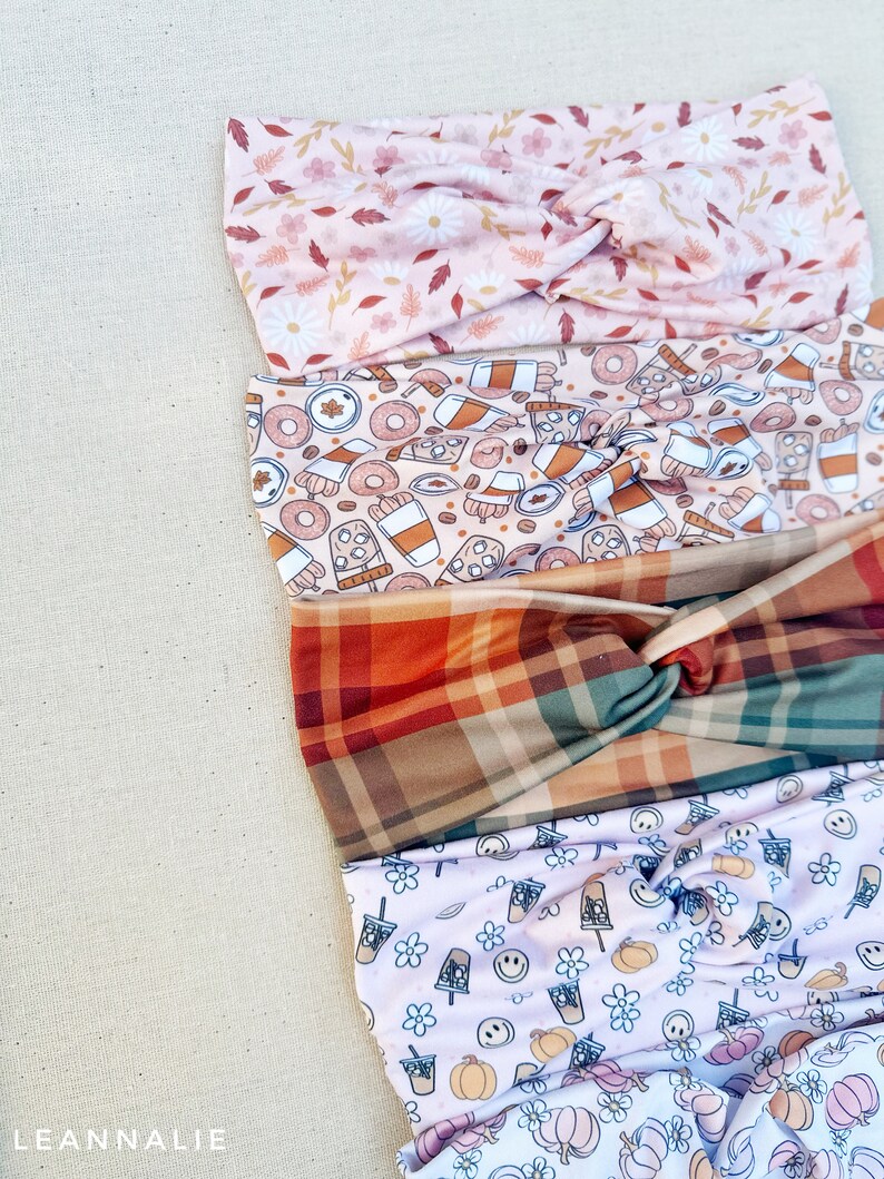 May include: Five different patterned headbands stacked on top of each other. The top headband is pink with white flowers and brown leaves. The second headband is pink with a brown and white donut, coffee cup, and pumpkin pattern. The third headband is a plaid pattern with red, green, and brown squares. The fourth headband is white with a light pink pumpkin, coffee cup, and smiley face pattern. The bottom headband is white with a pink pumpkin pattern.