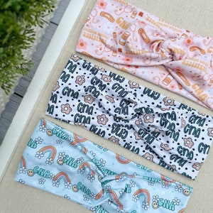 May include: Three colorful headbands with different patterns. The top headband is pink with a white background and features the text "certified nursing assistant" and medical symbols. The middle headband is white with a black background and features the text "cna" and small flowers. The bottom headband is light blue with a white background and features rainbows, smiley faces, and the text "cna".