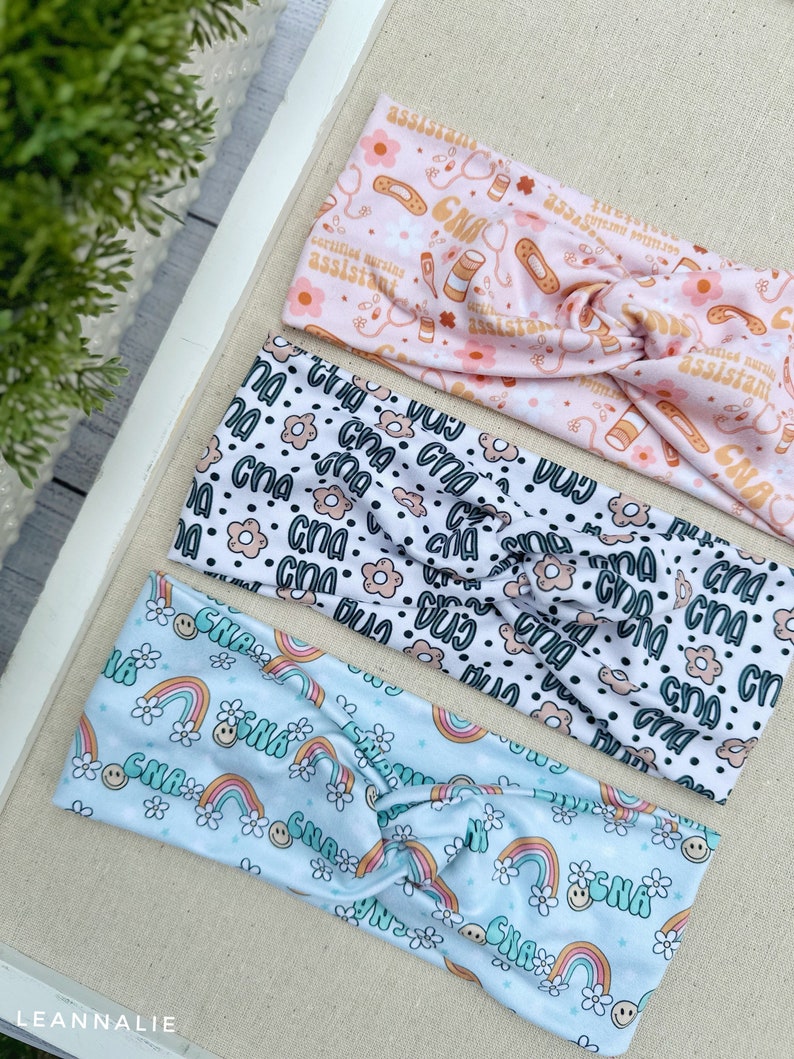 May include: Three colorful headbands with different patterns. The top headband is pink with a white background and features the text "certified nursing assistant" and medical symbols. The middle headband is white with a black background and features the text "cna" and small flowers. The bottom headband is light blue with a white background and features rainbows, smiley faces, and the text "cna".