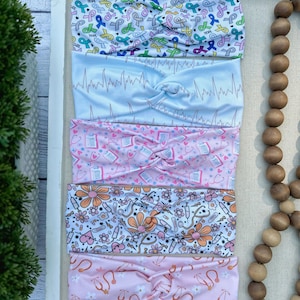 K&ouml;nnte beinhalten: Satz von f&uuml;nf bunten Stirnb&auml;ndern mit verschiedenen Mustern. Die Stirnb&auml;nder zeigen eine Vielzahl von Designs, darunter rosa Schleifen, EKG-Linien, medizinische Symbole und Blumen. Die Stirnb&auml;nder sind alle aus einem weichen, dehnbaren Stoff gefertigt.