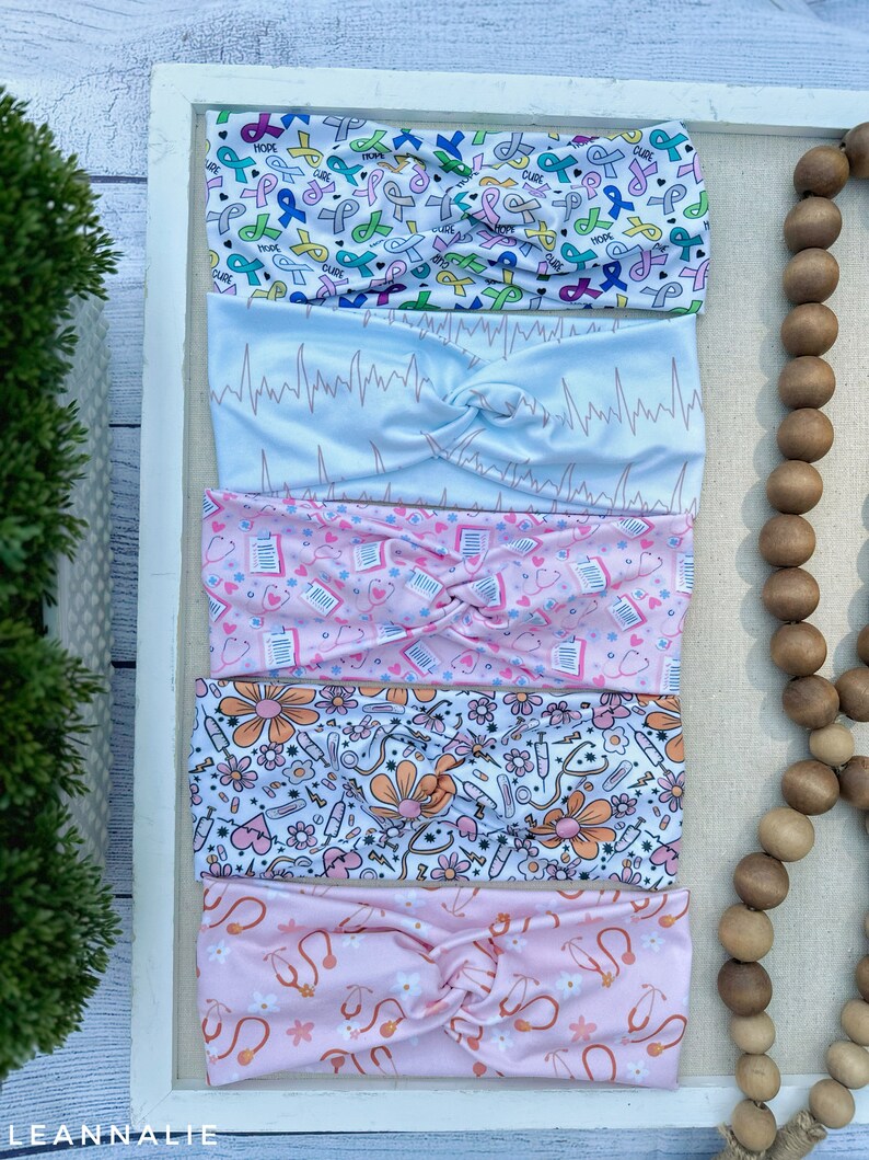 K&ouml;nnte beinhalten: Satz von f&uuml;nf bunten Stirnb&auml;ndern mit verschiedenen Mustern. Die Stirnb&auml;nder zeigen eine Vielzahl von Designs, darunter rosa Schleifen, EKG-Linien, medizinische Symbole und Blumen. Die Stirnb&auml;nder sind alle aus einem weichen, dehnbaren Stoff gefertigt.
