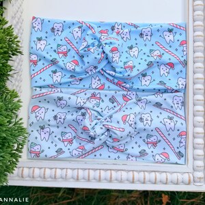 May include: Two light blue headbands with a repeating pattern of cartoon teeth wearing Santa hats, holding toothbrushes and candy canes. The headbands are twisted at the top. The headbands are displayed on a white framed surface.