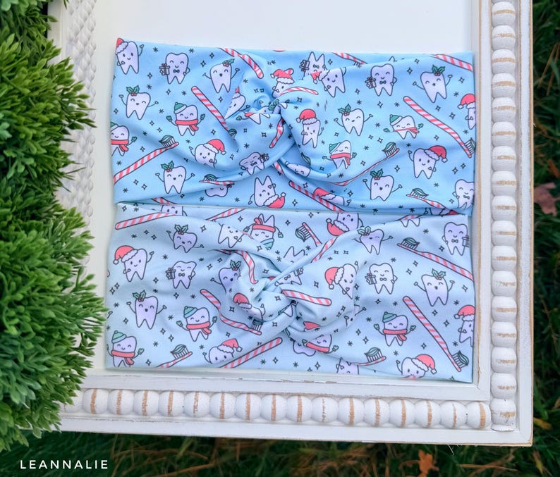 May include: Two light blue headbands with a repeating pattern of cartoon teeth wearing Santa hats, holding toothbrushes and candy canes. The headbands are twisted at the top. The headbands are displayed on a white framed surface.