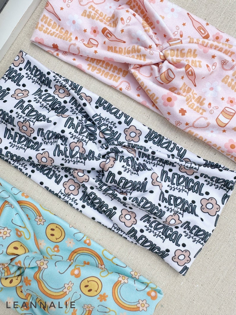 May include: Three different patterned headbands. The first headband is pink with a white background and features a repeating pattern of medical symbols and the text "Medical Assistant". The second headband is white with a black background and features a repeating pattern of black dots, pink flowers, and the text "Medical Assistant". The third headband is light blue with a white background and features a repeating pattern of rainbows, yellow smiley faces, and white flowers.