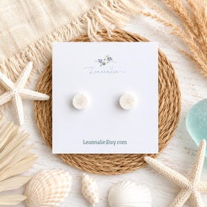 May include: A pair of round, off-white stud earrings featuring a delicate, embossed sand dollar pattern. The earrings are mounted on a white rectangular display card with the brand name Leannalie and the website Leannalie.Etsy.com printed on it.