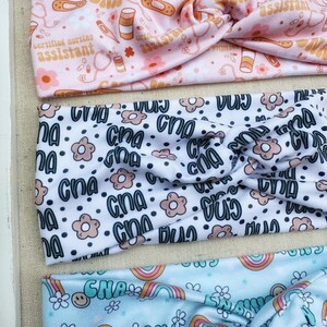 May include: Three colorful headbands with different patterns. The first headband is pink with a white background and features a repeating pattern of pills, bandages, stethoscopes, and the text "certified nursing assistant". The second headband is white with a black background and features a repeating pattern of the text "CNA" and small flowers. The third headband is light blue with a white background and features a repeating pattern of rainbows and the text "CNA".