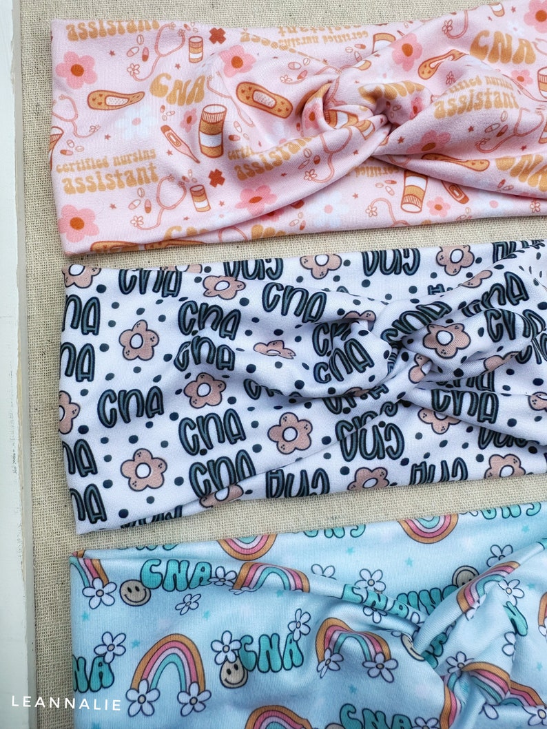 May include: Three colorful headbands with different patterns. The first headband is pink with a white background and features a repeating pattern of pills, bandages, stethoscopes, and the text "certified nursing assistant". The second headband is white with a black background and features a repeating pattern of the text "CNA" and small flowers. The third headband is light blue with a white background and features a repeating pattern of rainbows and the text "CNA".