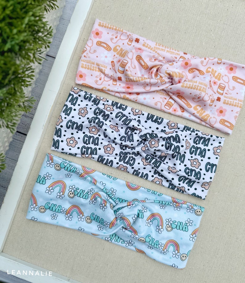 May include: Three colorful headbands with different patterns. The top headband is pink with a white pattern of medical symbols, the middle headband is white with a black and brown pattern of flowers and the text "CNA", and the bottom headband is light blue with a white pattern of rainbows and the text "CNA".