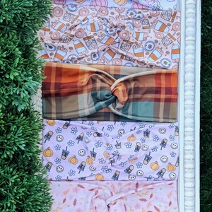May include: Five fabric headbands with twisted knots, featuring various patterns including pumpkins, coffee cups, plaid, smiley faces, and autumn leaves in shades of orange, pink, purple, and brown, arranged vertically on a white surface.