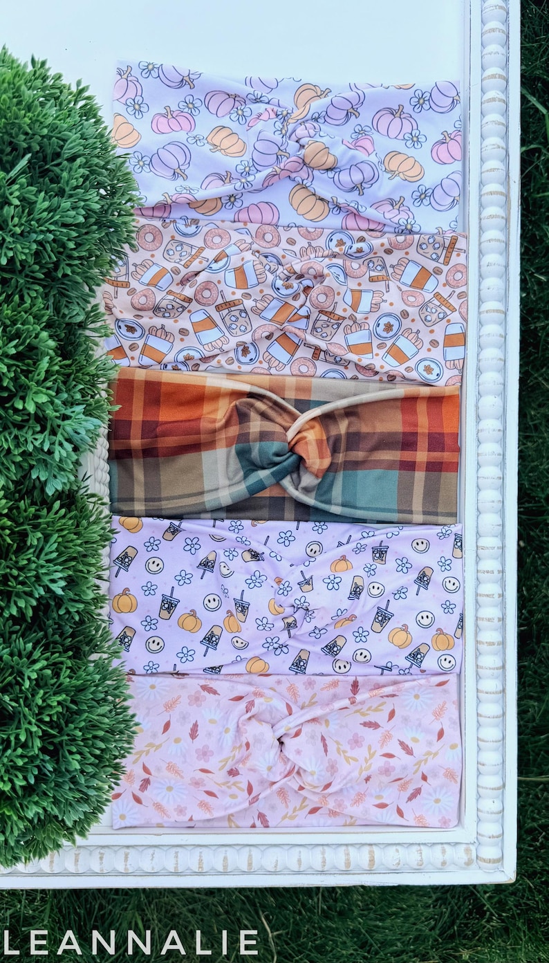 May include: Five fabric headbands with twisted knots, featuring various patterns including pumpkins, coffee cups, plaid, smiley faces, and autumn leaves in shades of orange, pink, purple, and brown, arranged vertically on a white surface.