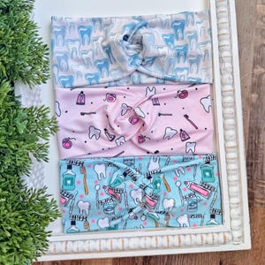 Könnte beinhalten: Drei Stoff-Haarbänder mit Dentalmustern. Das obere Haarband ist weiß mit blauen Zähnen, das mittlere ist rosa mit zahnärztlichen Werkzeugen und das untere ist türkis mit Dentalzubehör. Die Haarbänder werden in einem weiß gerahmten Tablett präsentiert.