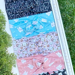 Könnte beinhalten: Sechs Stoff-Haarbänder mit Knoten-Design. Die Haarbänder zeigen verschiedene Muster, darunter Zähne, Sterne und Regenbogen, in Farben wie Rosa, Schwarz und Blau. Sie werden auf einem weißen Tablett präsentiert.