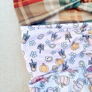 May include: Three fabric headbands with different patterns. The top headband is a plaid pattern in shades of brown, orange, and green. The middle headband has a pink background with a white pattern of pumpkins, coffee cups, and smiley faces. The bottom headband has a white background with a pink and orange pumpkin pattern.