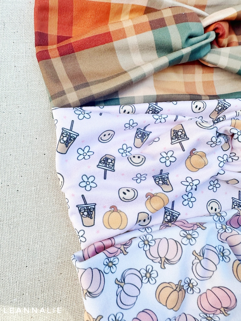 May include: Three fabric headbands with different patterns. The top headband is a plaid pattern in shades of brown, orange, and green. The middle headband has a pink background with a white pattern of pumpkins, coffee cups, and smiley faces. The bottom headband has a white background with a pink and orange pumpkin pattern.