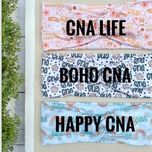 May include: Three different headbands with a variety of patterns. The first headband is pink with a white pattern of medical symbols and the text "CNA LIFE". The second headband is white with a black pattern of flowers and the text "BOHO CNA". The third headband is light blue with a white pattern of rainbows and the text "HAPPY CNA".
