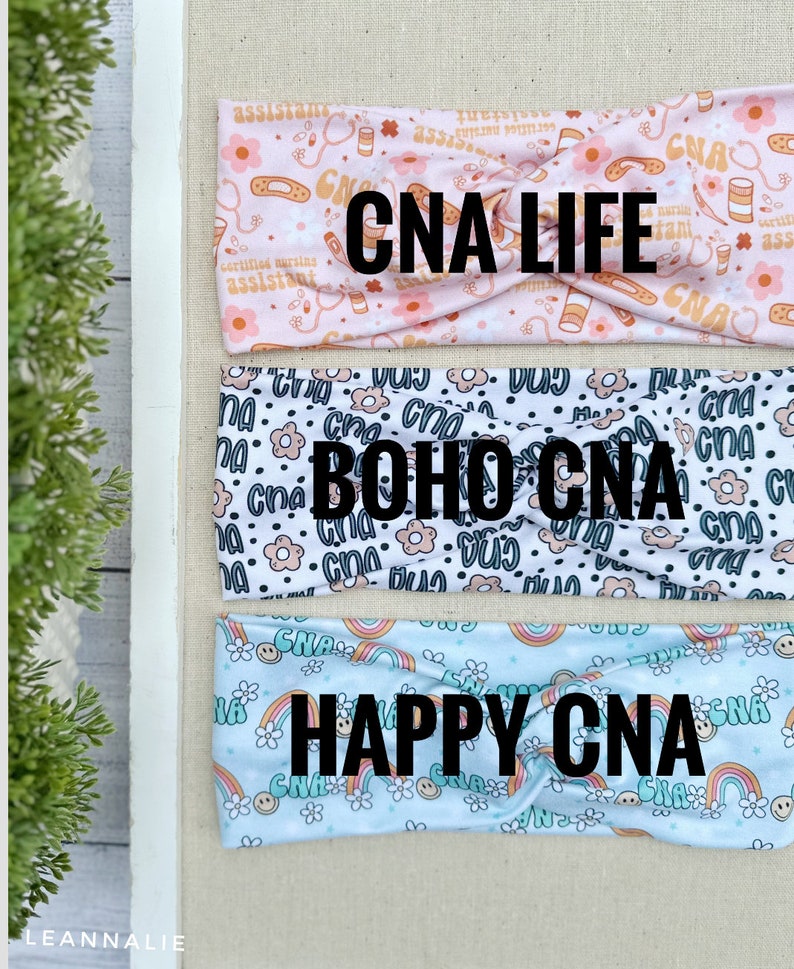 May include: Three different headbands with a variety of patterns. The first headband is pink with a white pattern of medical symbols and the text "CNA LIFE". The second headband is white with a black pattern of flowers and the text "BOHO CNA". The third headband is light blue with a white pattern of rainbows and the text "HAPPY CNA".