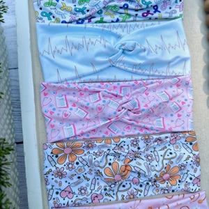 K&ouml;nnte beinhalten: F&uuml;nf verschiedene gemusterte Stirnb&auml;nder, alle mit einem verdrehten Knoten in der Mitte. Das obere Stirnband ist wei&szlig; mit rosa, blauen und gr&uuml;nen Banddesigns und den Worten "HOPE" und "CURE". Das zweite Stirnband ist hellblau mit einem wei&szlig;en EKG-Muster. Das dritte Stirnband ist rosa mit einem wei&szlig;en Muster aus medizinischen Symbolen, darunter Stethoskope, Pillen und Verb&auml;nde. Das vierte Stirnband ist wei&szlig; mit einem rosa und orangefarbenen Muster aus Blumen, Spritzen und Stethoskopen. Das untere Stirnband ist rosa mit einem wei&szlig;en Muster aus Blumen und Stethoskopen.