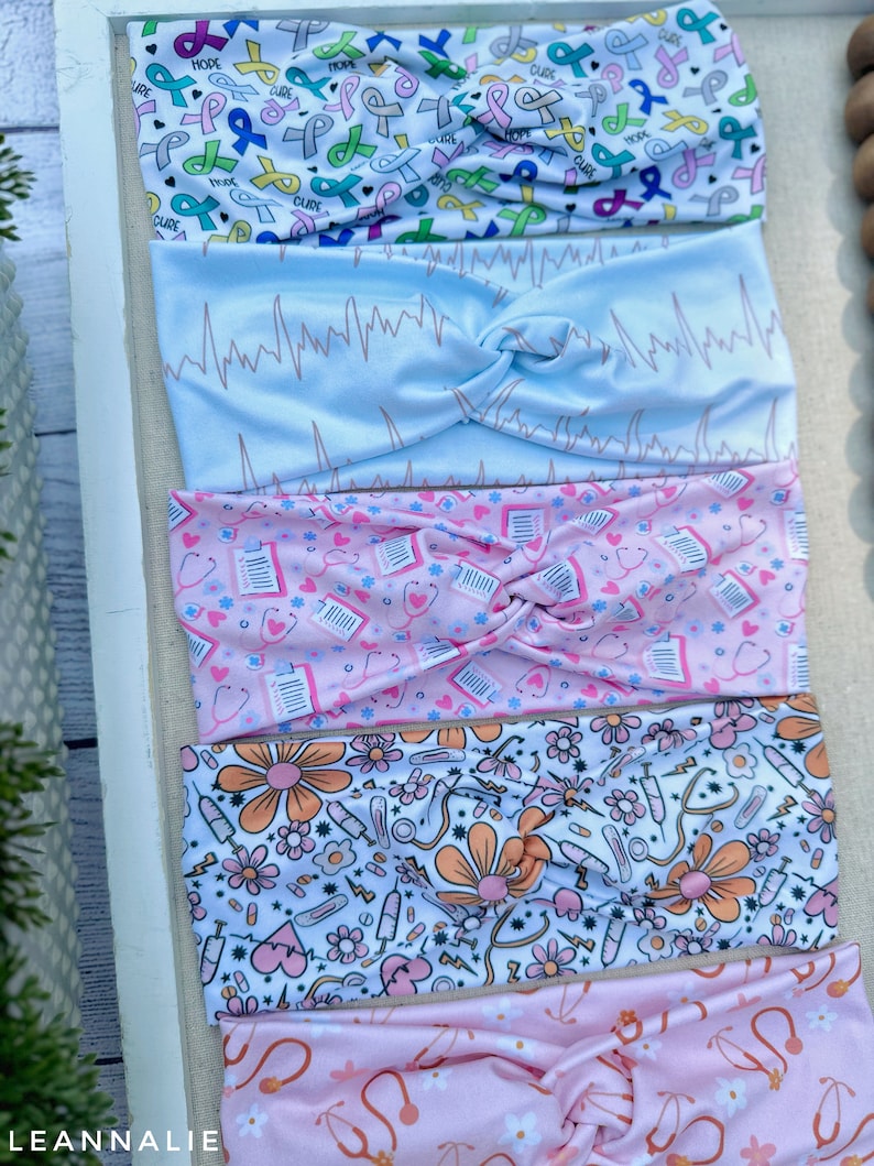 K&ouml;nnte beinhalten: F&uuml;nf verschiedene gemusterte Stirnb&auml;nder, alle mit einem verdrehten Knoten in der Mitte. Das obere Stirnband ist wei&szlig; mit rosa, blauen und gr&uuml;nen Banddesigns und den Worten "HOPE" und "CURE". Das zweite Stirnband ist hellblau mit einem wei&szlig;en EKG-Muster. Das dritte Stirnband ist rosa mit einem wei&szlig;en Muster aus medizinischen Symbolen, darunter Stethoskope, Pillen und Verb&auml;nde. Das vierte Stirnband ist wei&szlig; mit einem rosa und orangefarbenen Muster aus Blumen, Spritzen und Stethoskopen. Das untere Stirnband ist rosa mit einem wei&szlig;en Muster aus Blumen und Stethoskopen.