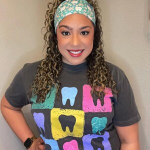May include: A charcoal gray short-sleeve t-shirt featuring a grid of colorful, distressed-style squares, each containing a solid black tooth icon. The shirt is paired with a light blue headband decorated with a repeating pattern of small white teeth and flowers.