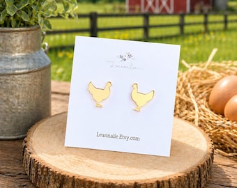 May include: Gold-coloured chicken-shaped stud earrings on a white card, displayed on a wooden slice. The card has the text "Leannalie" and "Leannalie.Etsy.com". A red barn, green grass, and blue sky form the background.