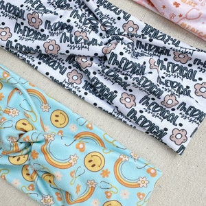 May include: Three fabric headbands with different patterns. The first headband is white with black text that says "Medical Assistant". The second headband is light blue with a pattern of rainbows, smiley faces, and flowers. The third headband is pink with a pattern of medical symbols and flowers.