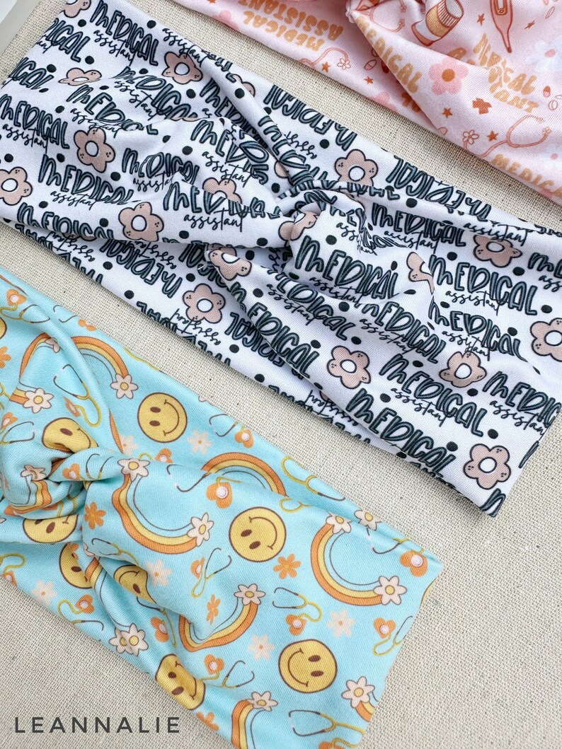 May include: Three fabric headbands with different patterns. The first headband is white with black text that says "Medical Assistant". The second headband is light blue with a pattern of rainbows, smiley faces, and flowers. The third headband is pink with a pattern of medical symbols and flowers.