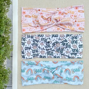 May include: Three colorful headbands with different patterns. The top headband is pink with a white background and features a repeating pattern of the text "CNA", flowers, and medical symbols. The middle headband is white with a black background and features a repeating pattern of the text "CNA" and flowers. The bottom headband is light blue with a white background and features a repeating pattern of the text "CNA", rainbows, and smiley faces.