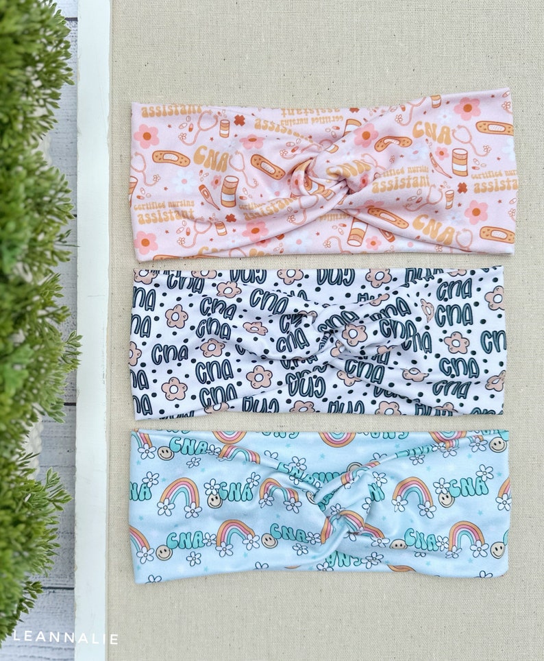 May include: Three colorful headbands with different patterns. The top headband is pink with a white background and features a repeating pattern of the text "CNA", flowers, and medical symbols. The middle headband is white with a black background and features a repeating pattern of the text "CNA" and flowers. The bottom headband is light blue with a white background and features a repeating pattern of the text "CNA", rainbows, and smiley faces.