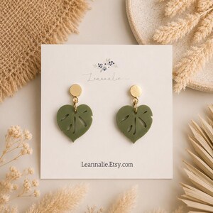 May include: A pair of dangle earrings featuring matte olive green heart-shaped leaf charms with cut-out details, suspended from small gold-tone circular studs. The earrings are displayed on a white card printed with the text Leannalie and Leannalie.Etsy.com.