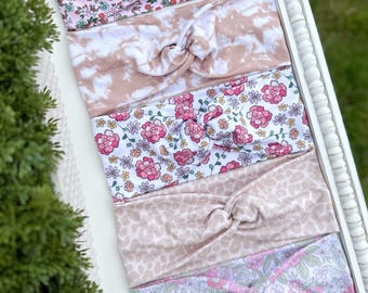 May include: A collection of six knotted headbands in various floral and patterned designs. The headbands feature colors like pink, white, and tan. The headbands are displayed on a white tray with a decorative border. The brand name "LEANNALIE" is visible in the bottom left corner.