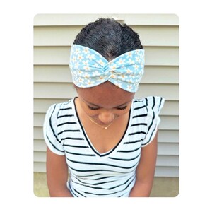 May include: A light blue fabric headband featuring a repeating white daisy floral pattern with a central twist detail. The headband is worn across the forehead, showcasing a soft textile construction with a consistent print throughout the fabric.
