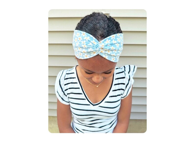 May include: A light blue fabric headband featuring a repeating white daisy floral pattern with a central twist detail. The headband is worn across the forehead, showcasing a soft textile construction with a consistent print throughout the fabric.
