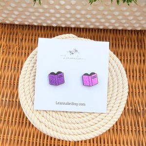 May include: A pair of purple book-shaped earrings with glitter accents, displayed on a white card with the text "Leannalie Etsy.com". The earrings are set on a cream-colored coaster, with a woven wicker background.