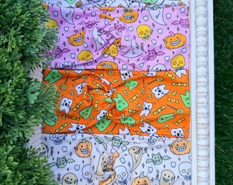 May include: Four fabric headbands with twisted front details, featuring repeating patterns of Halloween-themed icons including ghosts, smiling pumpkins, skulls, bats, candy, and teeth in white, orange, pink, and light green color palettes.