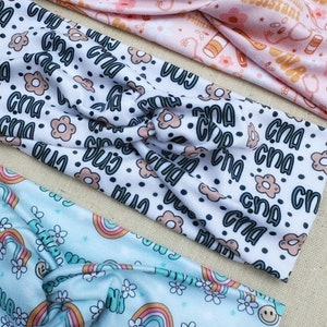 May include: Three different patterned headbands. The first headband is pink with a white pattern of pills, bandages, and the text "certified nursing assistant". The second headband is white with a black polka dot pattern and the text "CNA" repeated in a variety of fonts and colors. The third headband is light blue with a white pattern of rainbows, flowers, and the text "CNA" in a retro font.