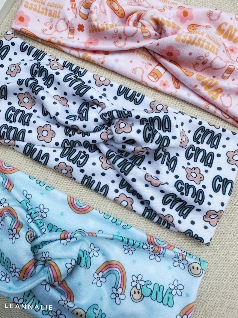 May include: Three different patterned headbands. The first headband is pink with a white pattern of pills, bandages, and the text "certified nursing assistant". The second headband is white with a black polka dot pattern and the text "CNA" repeated in a variety of fonts and colors. The third headband is light blue with a white pattern of rainbows, flowers, and the text "CNA" in a retro font.