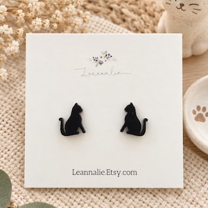 Può includere: Un paio di orecchini a lobo a forma di gatto nero presentati su un cartoncino rettangolare bianco. Il cartoncino presenta una piccola grafica floreale, il nome del marchio Leannalie in corsivo e l'indirizzo Leannalie.Etsy.com.