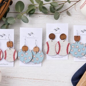 May include: Baseball-themed earrings with wooden accents. The earrings feature baseball designs in white and light blue, with red stitching details. A baseball glove, baseball, and bat are also in the image.