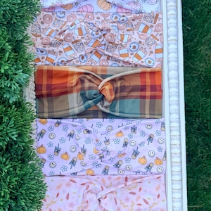 May include: Five fabric headbands with twisted knots, featuring various patterns including orange and purple pumpkins, coffee cups with donuts, a multi-colored plaid, smiley faces with pumpkins, and a light pink floral print with autumn leaves.