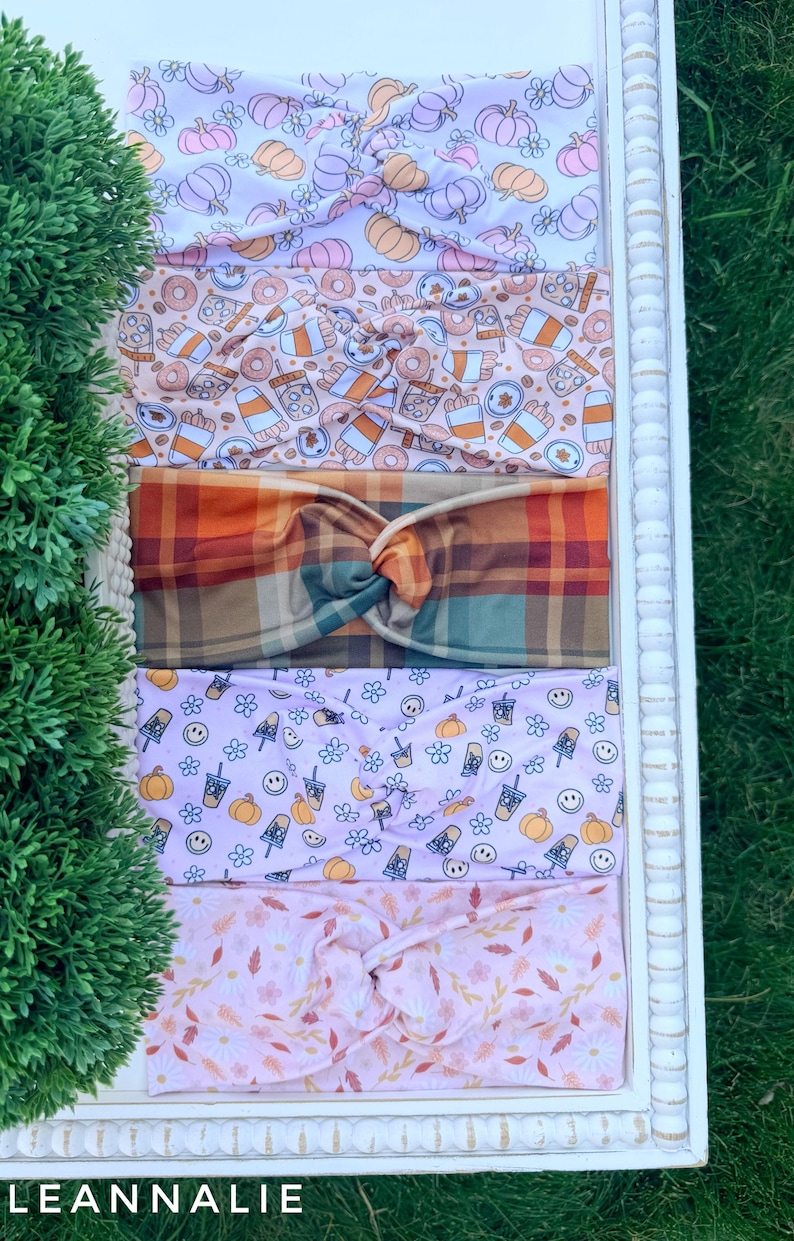 May include: Five fabric headbands with twisted knots, featuring various patterns including orange and purple pumpkins, coffee cups with donuts, a multi-colored plaid, smiley faces with pumpkins, and a light pink floral print with autumn leaves.