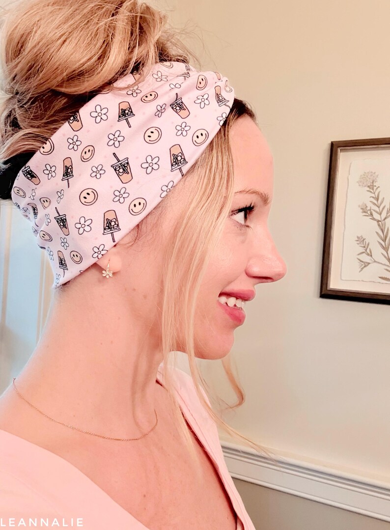 May include: A light pink headband featuring a repeating pattern of iced coffee cups, smiling faces, and small flowers in black and brown outlines. The fabric accessory is worn around the head, displaying a wide, knotted design with a soft finish.