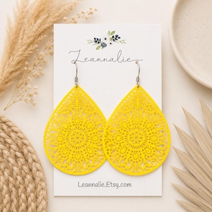 May include: A pair of bright yellow teardrop-shaped earrings featuring an intricate laser-cut mandala pattern. The earrings hang from silver-toned metal hooks and are displayed on a white card printed with the text Leannalie and Leannalie.Etsy.com.