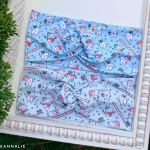 May include: Two light blue headbands with a repeating pattern of cartoon teeth wearing Santa hats and scarves, toothbrushes, and candy canes. The headbands are twisted at the center and are displayed on a white tray.