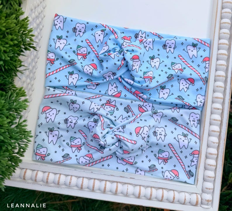 May include: Two light blue headbands with a repeating pattern of cartoon teeth wearing Santa hats and scarves, toothbrushes, and candy canes. The headbands are twisted at the center and are displayed on a white tray.