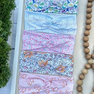 K&ouml;nnte beinhalten: Vier bunte Stirnb&auml;nder mit verschiedenen Mustern. Das obere Stirnband hat einen wei&szlig;en Hintergrund mit rosa, blauen und gr&uuml;nen B&auml;ndern mit den Worten "Hope", "Love" und "Cure". Das zweite Stirnband hat einen hellblauen Hintergrund mit einem wei&szlig;en EKG-Muster. Das dritte Stirnband hat einen rosa Hintergrund mit einem wei&szlig;en Muster aus Stethoskopen, Herzen und Pillen. Das vierte Stirnband hat einen wei&szlig;en Hintergrund mit einem rosa Muster aus Blumen, Stethoskopen und Pillen.