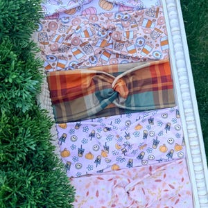 May include: Five fabric headbands with twisted knots, featuring various patterns including orange and purple pumpkins, coffee cups and donuts, a multi-colored plaid, smiley faces with pumpkins, and a light pink floral print with autumn leaves.