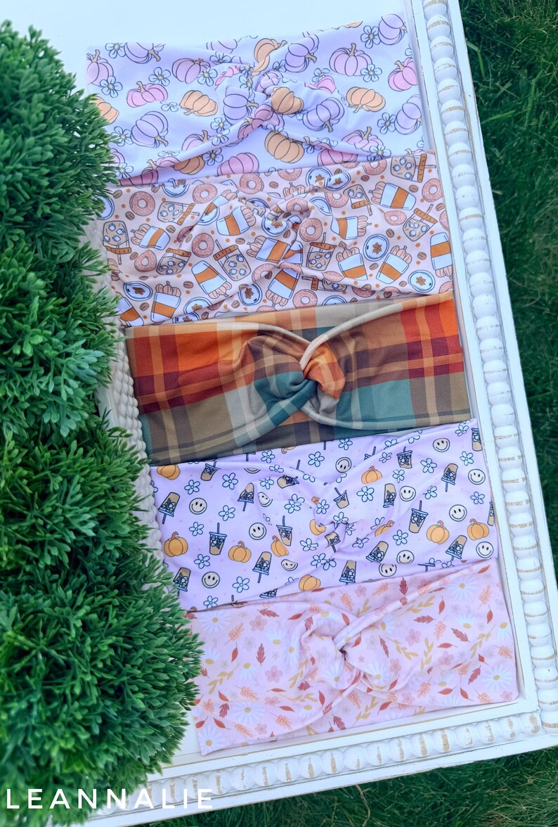 May include: Five fabric headbands with twisted knots, featuring various patterns including orange and purple pumpkins, coffee cups and donuts, a multi-colored plaid, smiley faces with pumpkins, and a light pink floral print with autumn leaves.