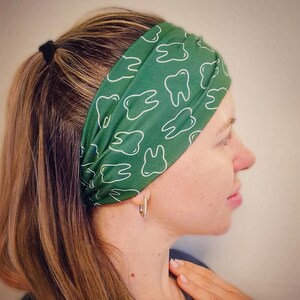 May include: A wide fabric headband in a deep forest green color featuring a repeating white line art pattern of human teeth. The headband is worn across the forehead and is made of a smooth, flexible material that fits snugly around the head.